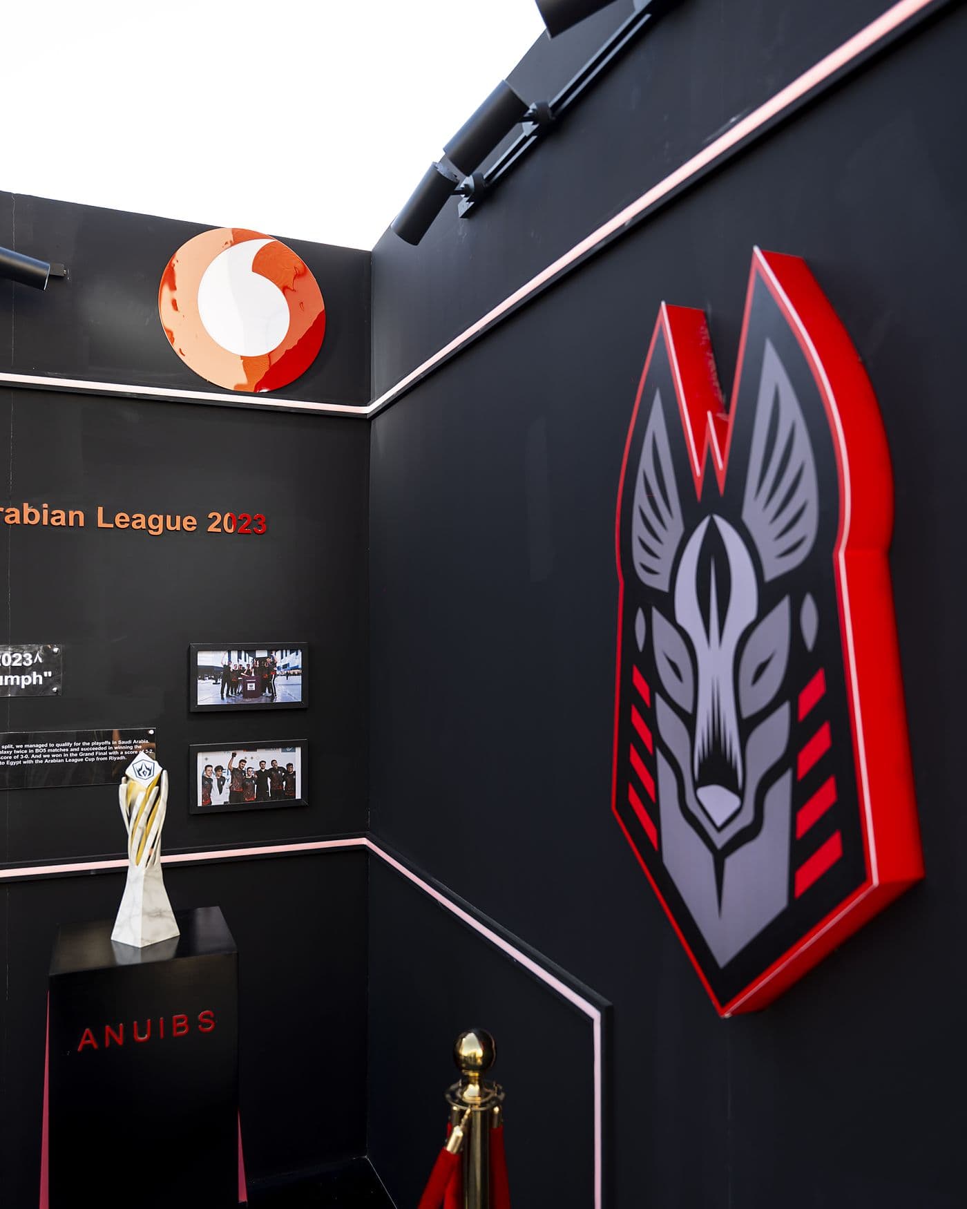 Black trophy room with a red-edged Anubis crest wall, Vodafone logo and the league trophy on a plinth